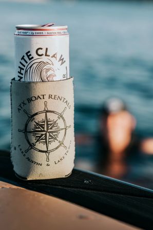 Tall white seltzer can tucked in a nautical compass koozie on a boat railing with sunlit lake water and a blurred swimmer in the background — summer boat life.