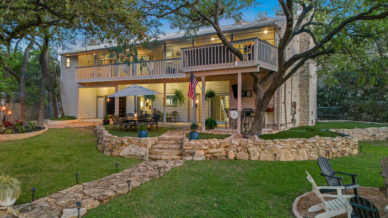 Inviting dusk backyard of a two-story home with an upper balcony and covered lower patio, stone retaining walls and winding flagstone path, outdoor dining set under an umbrella, Adirondack chairs, mature oak trees and landscaped lawn.