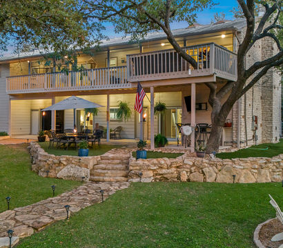 Inviting dusk backyard of a two-story home with an upper balcony and covered lower patio, stone retaining walls and winding flagstone path, outdoor dining set under an umbrella, Adirondack chairs, mature oak trees and landscaped lawn.