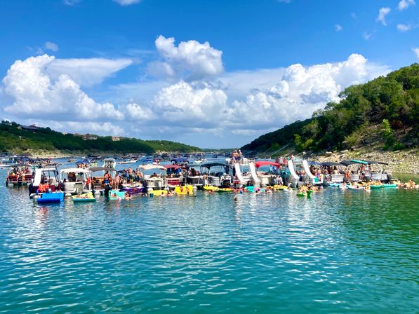 Summer boat party: crowded flotilla of colorful pontoon boats with slides and swimmers anchored in a sunny lake cove, surrounded by green hills and a blue sky dotted with puffy clouds.