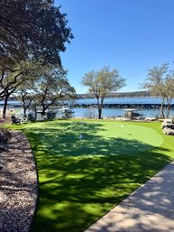 Sunlit lakefront putting green with small flags, Adirondack chairs under oak trees, a marina of covered boat slips and a calm blue lake under a clear sky.