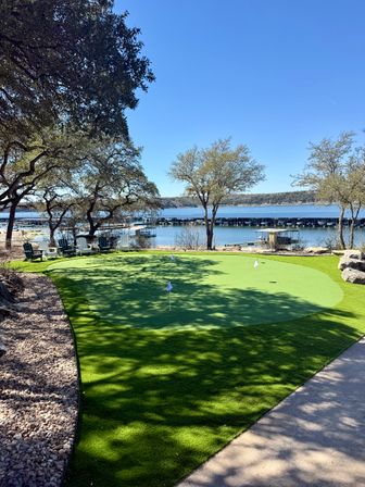 Sunlit lakefront putting green with small flags, Adirondack chairs under oak trees, a marina of covered boat slips and a calm blue lake under a clear sky.