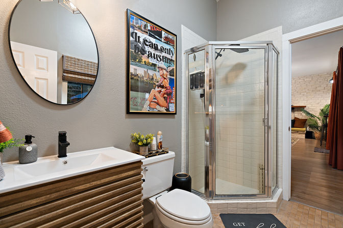 Modern apartment bathroom with corner glass shower, slatted wood vanity, round mirror, toilet and framed retro poster