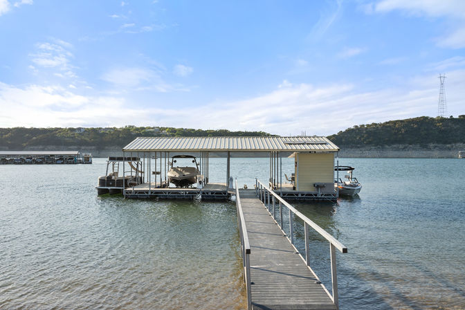Wooden walkway leading to a covered boat dock and small yellow boathouse with two boats on a calm lake, blue sky and tree‑covered hills in the background.