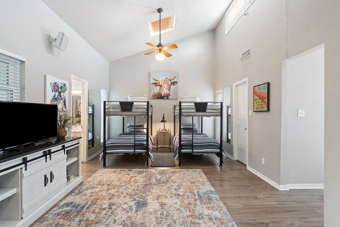Bright modern bunk bedroom with two black metal twin-over-twin bunk beds in striped linens, central nightstand and lamp under a colorful cow painting, ceiling fan with skylight, TV console, patterned area rug and wood-look floors.