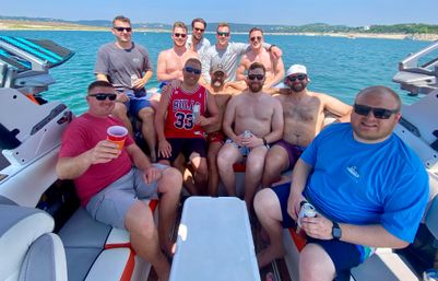 Group of men enjoying a summer boat party on clear blue water, lounging in a motorboat with drinks and a rocky shoreline in the background