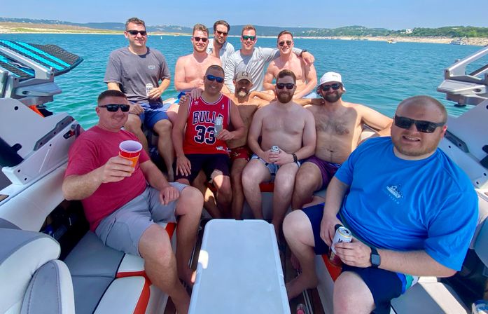 Group of men enjoying a summer boat party on clear blue water, lounging in a motorboat with drinks and a rocky shoreline in the background