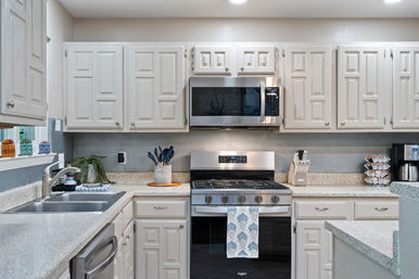 Bright modern white kitchen with paneled cabinets, speckled countertops, stainless steel gas range and over-the-range microwave, double sink and faucet, utensil jar and knife block, coffee maker with K‑cup carousel, and a patterned dish towel.