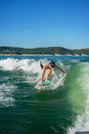 Wakesurfer crouching low on a turquoise board, carving a green wake near a hilly lakeshore under a clear blue summer sky — action-packed water sports scene.