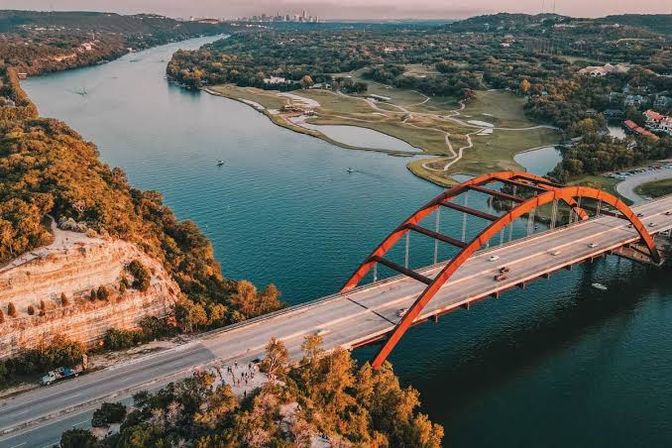 Aerial view of Austin’s red-arched Pennybacker (360) Bridge spanning Lake Austin at golden hour, with limestone cliffs, green parkland, and a distant skyline.