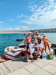Group of friends posing on a dock beside a red-and-white wakeboard boat at a sunny reservoir with blue skies and a distant concrete dam — fun summer lake day.