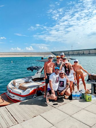 Group of friends posing on a dock beside a red-and-white wakeboard boat at a sunny reservoir with blue skies and a distant concrete dam — fun summer lake day.