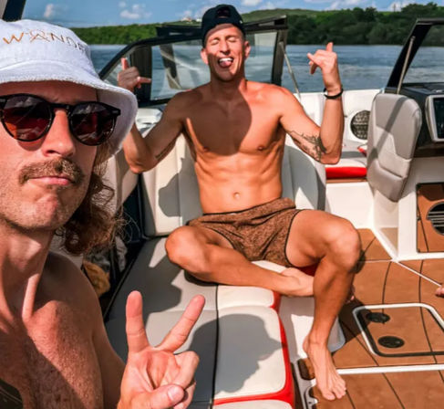 Two shirtless friends on a speedboat at a sunny lake—one in a bucket hat and sunglasses flashing a peace sign, the other cross-legged, tongue out and making playful hand gestures.
