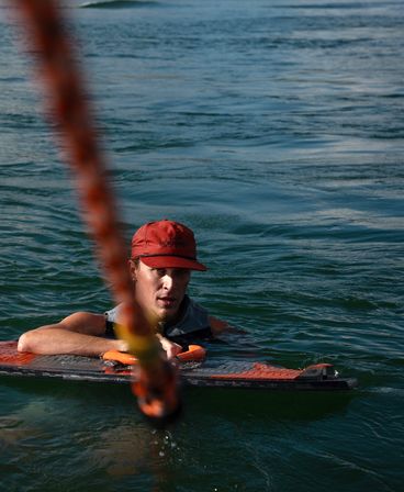 Wakeboarder in a red cap floating in green-blue open water, gripping an orange tow-rope handle with the board beneath — close-up watersports action shot