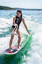 Smiling woman wakesurfing on a colorful board behind a boat on a sunny lake, carving a green wake
