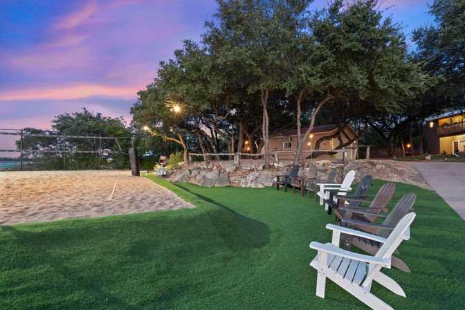 Sunset outdoor recreation area with a sand volleyball court, row of Adirondack chairs on green turf beneath oak trees strung with warm lights.
