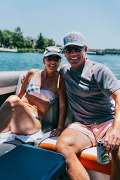 Smiling couple in sunglasses and caps relaxing on a boat at a sunny lake — woman in a tie-dye bikini, man holding a canned drink with shoreline and trees in the background.