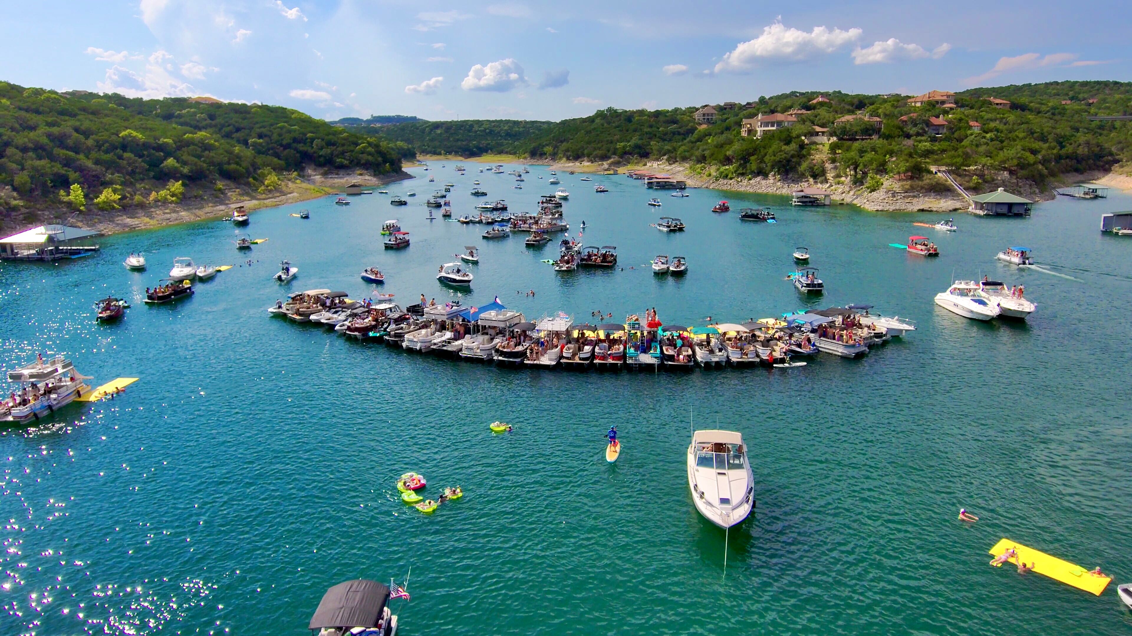 Aerial lake view of a busy boat party: dozens of pontoons and recreational boats linked into a floating platform, with inflatables and paddleboards on turquoise water and tree‑lined hills and shoreline homes in the background.