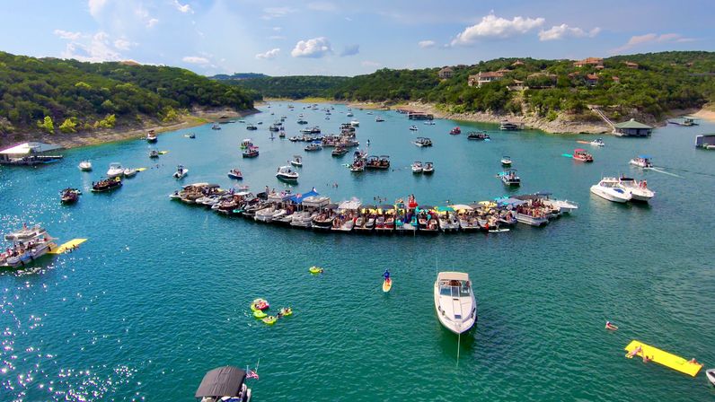 Aerial lake view of a busy boat party: dozens of pontoons and recreational boats linked into a floating platform, with inflatables and paddleboards on turquoise water and tree‑lined hills and shoreline homes in the background.
