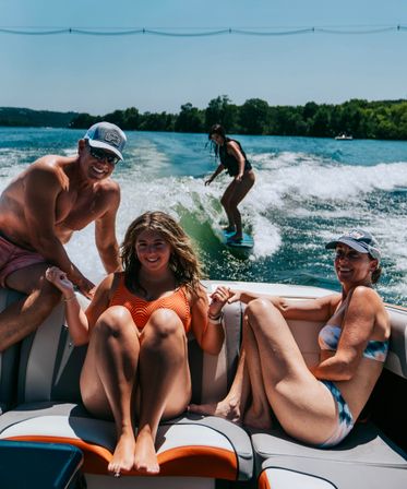 Sunny lake day: three smiling people lounging on a motorboat in swimsuits while a woman wakesurfs the boat's wake with a tree-lined shoreline in the background.