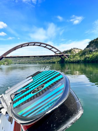 Turquoise-striped watersports board on a boat with a calm river, tree-lined banks and a rust-colored arched bridge under a bright blue sky