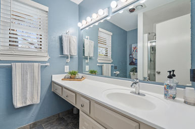 Bright blue bathroom with white vanity and integrated sink, chrome faucet, globe-lit mirror, striped towels, succulent tray and glass shower
