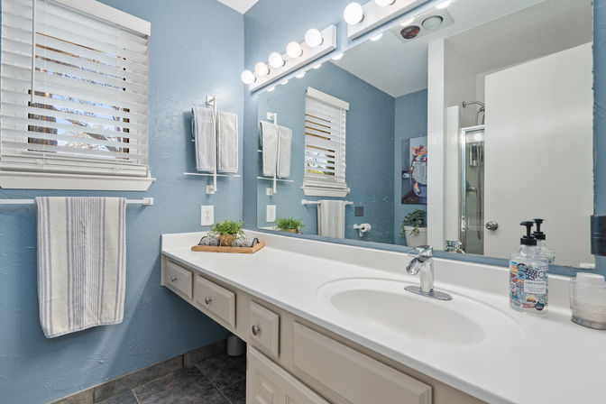Bright blue bathroom with white vanity and integrated sink, chrome faucet, globe-lit mirror, striped towels, succulent tray and glass shower