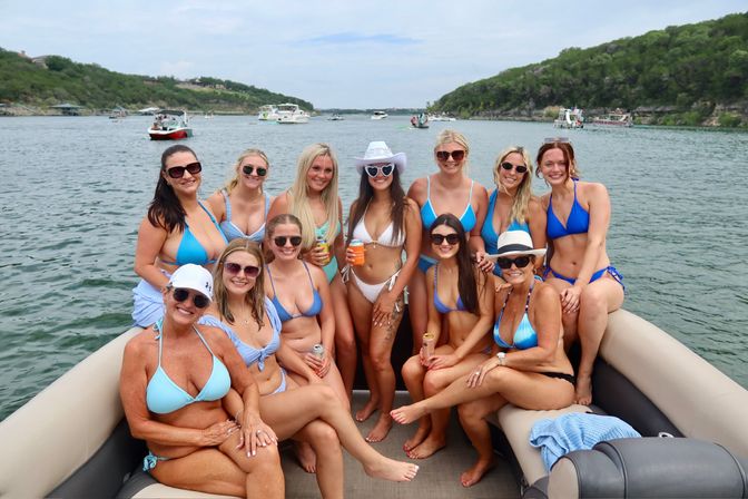 Group of women in colorful bikinis posing and smiling on a pontoon boat on a sunny lake, with other boats and a tree-lined shoreline in the background