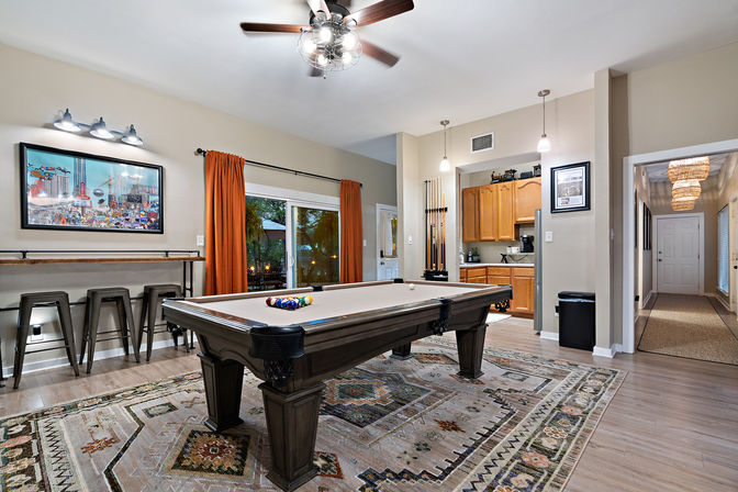 Inviting open-plan billiards room with wooden pool table on a patterned rug, ceiling fan and pendant lights, bar stools at a counter, sliding glass door with orange curtains, framed wall art, and a kitchenette with wall-mounted pool cues.