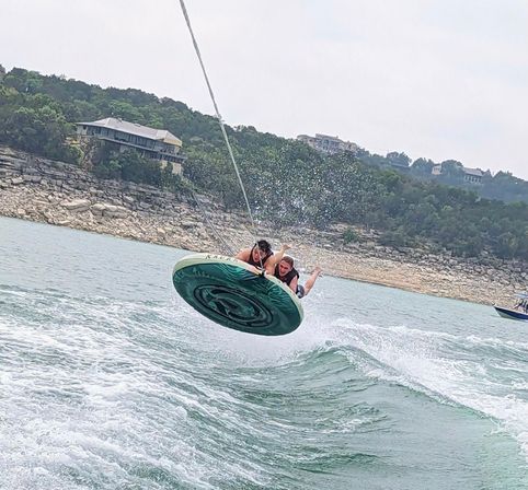 Two people laughing on a green inflatable tow tube as it soars over choppy lake water, splashing toward a rocky shoreline with forested hills and lakeside homes in the background.