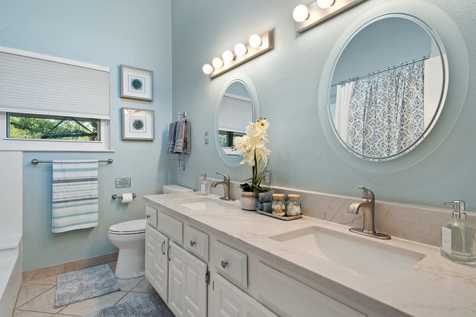 Bright coastal‑inspired bathroom with light blue walls, marble double-sink vanity, round mirrors, white orchid centerpiece and patterned shower curtain