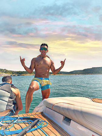 Shirtless man in colorful swim trunks and mirrored sunglasses striking a playful pose on a motorboat swim platform, friend in a life vest nearby, calm lake waters and rolling hills under a pastel sunset — summer boating scene.