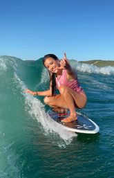 Smiling surfer in a pink vest crouched on a surfboard riding a green ocean wave, flashing a shaka hand sign with coastal hills and clear blue sky in the background.