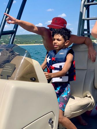 Young boy wearing a star-print life jacket steers a boat as an adult in a red cap points toward the scenic lake shoreline on a sunny summer day.