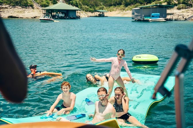 Kids laughing and playing on a turquoise inflatable mat in a sunny lake, wearing swimsuits and goggles with docks, houseboats, and a green inner tube in the background