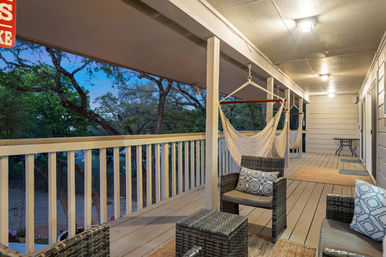 Cozy covered wooden porch at dusk with hanging hammock chairs, wicker seating and patterned pillows, warm ceiling lights and tree-lined yard view
