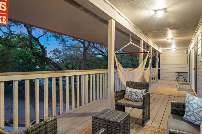 Cozy covered wooden porch at dusk with hanging hammock chairs, wicker seating and patterned pillows, warm ceiling lights and tree-lined yard view