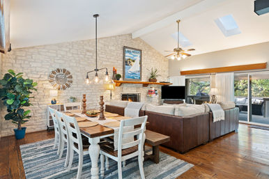 Sunlit open-concept living and dining room with a farmhouse wood table and white chairs on a striped rug, leather sectional facing a stone fireplace under skylights, sailboat artwork, and sliding glass doors to a tree-lined deck.