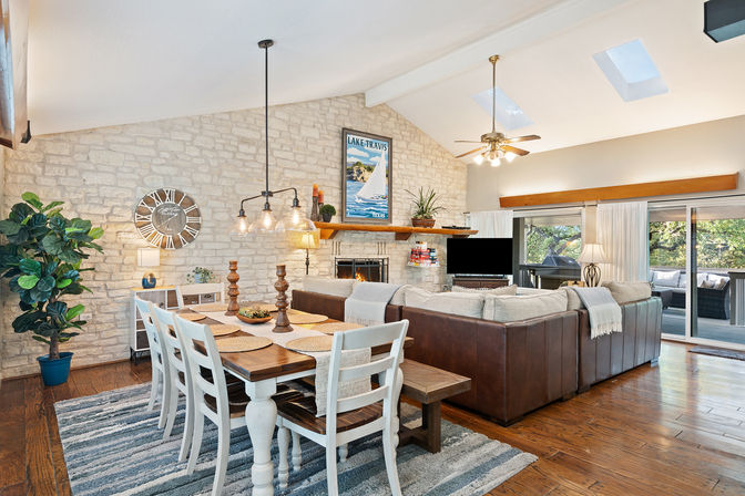 Sunlit open-concept living and dining room with a farmhouse wood table and white chairs on a striped rug, leather sectional facing a stone fireplace under skylights, sailboat artwork, and sliding glass doors to a tree-lined deck.