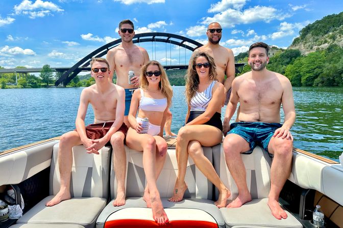 Six friends in swimwear relaxing on a motorboat on a sunny river with an arched bridge and green cliffs in the background