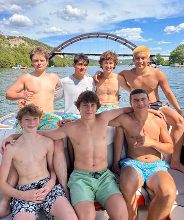 Seven teenage friends on a motorboat enjoying a sunny summer lake day, shirtless in swim trunks and smiling with an arched bridge and tree-lined shoreline in the background.