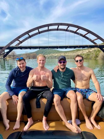 Four friends smiling and posing on a boat beneath a large arched steel bridge over a calm lake, wooded hills and blue sky in the background