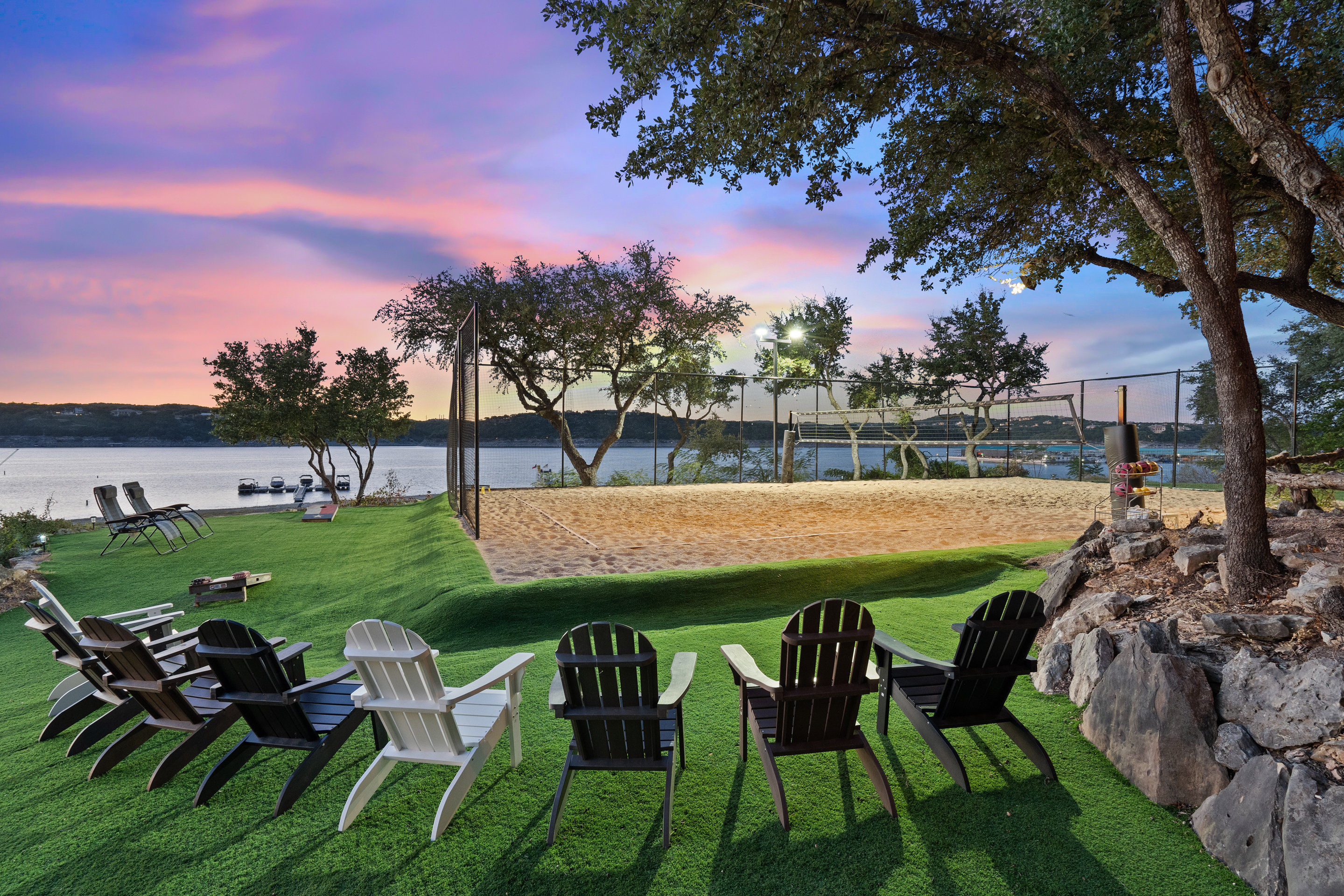 Lakefront sunset with Adirondack chairs on a manicured green lawn facing calm water, a sandy beach volleyball court with lights, docked boats and silhouetted trees.