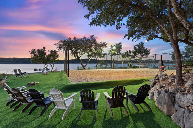 Lakefront sunset with Adirondack chairs on a manicured green lawn facing calm water, a sandy beach volleyball court with lights, docked boats and silhouetted trees.