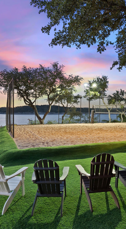 Lakefront sunset with Adirondack chairs on a manicured green lawn facing calm water, a sandy beach volleyball court with lights, docked boats and silhouetted trees.