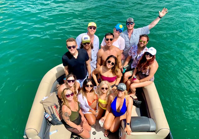 Group of friends on a pontoon boat in turquoise water, smiling in swimsuits and sunglasses and enjoying a sunny summer boating day with drinks.