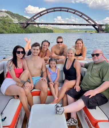 Eight people, including a young girl, smiling on a motorboat under a large arched bridge over a tree-lined river on a sunny summer day.