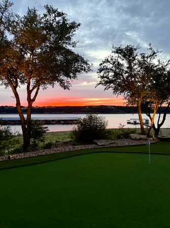 Vibrant orange-pink lake sunset framed by silhouetted trees, marina docks along the far shore and a manicured putting green in the foreground.