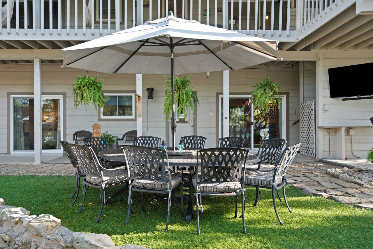 Backyard patio with round wrought-iron dining table and cushioned chairs under a large beige umbrella on green lawn, hanging fern planters and a two-story deck with sliding doors in the background.