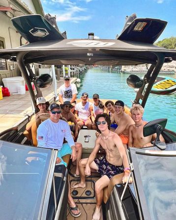 Group of friends relaxing on a modern wakeboard boat at a sunny marina, lounging on tan seats with turquoise water, docks and a paddleboard visible in the background, casual summer attire and sunglasses.